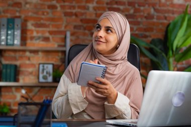 Orta Doğulu bej tesettürlü Müslüman kadın çatı katında masa başında çalışıyor. Kadın fikirlerini ya da bilgilerini kalemle deftere yazıyor..
