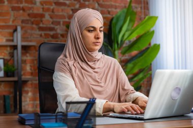 Orta Doğulu bej tesettürlü Müslüman kadın çatı katında masa başında çalışıyor. Kadın dizüstü bilgisayarda klavye kullanıyor..