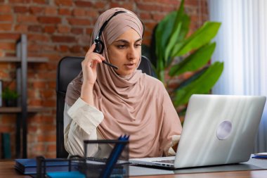 Orta Doğulu bej tesettürlü Müslüman kadın çatı katında masa başında çalışıyor. Çağrı merkezindeki kadın bir müşteriyle iletişim kuruyor ve destek ve yardım sağlıyor..