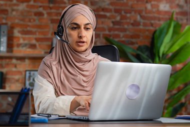 Orta Doğulu bej tesettürlü Müslüman kadın çatı katında masa başında çalışıyor. Çağrı merkezindeki kadın bir müşteriyle iletişim kuruyor ve destek ve yardım sağlıyor..