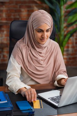 Odaklanmış, bej tesettürlü, tavan arası tarzı bir ofiste çalışan, elinde kredi kartı olan ve internetten alışveriş yapmak için dizüstü bilgisayar kullanan Ortadoğulu Müslüman bir kadın..