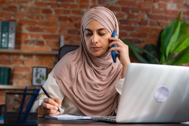 Orta Doğulu bej tesettürlü Müslüman kadın çatı katında masa başında çalışıyor. Kadın telefonda konuşuyor ve bir deftere bilgi yazıyor..