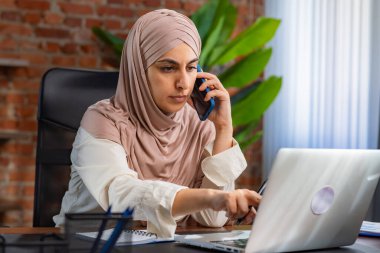 Orta Doğulu bej tesettürlü Müslüman kadın çatı katında masa başında çalışıyor. Kadın akıllı telefonuyla konuşuyor ve bilgisayarındaki bilgileri izliyor..