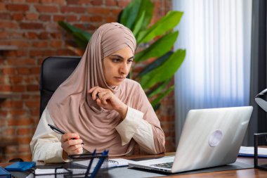Orta Doğulu bej tesettürlü Müslüman kadın çatı katında masa başında çalışıyor. Kadın dizüstü bilgisayarındaki bilgilere bakıyor ve kalemle deftere yazıyor..