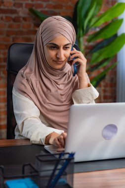 Orta Doğulu bej tesettürlü Müslüman kadın çatı katında masa başında çalışıyor. Kadın akıllı telefonuyla konuşuyor ve bilgisayarındaki bilgileri izliyor..