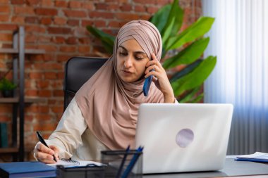 Orta Doğulu bej tesettürlü Müslüman kadın çatı katında masa başında çalışıyor. Kadın telefonda konuşuyor ve bir deftere bilgi yazıyor..