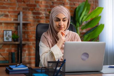 Orta Doğulu bej tesettürlü Müslüman kadın çatı katında masa başında çalışıyor. Kadın laptopuyla çalışıyor ve bir şeyler düşünüyor..