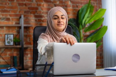 Orta Doğulu bej tesettürlü Müslüman kadın çatı katında masa başında çalışıyor. Kadın dizüstü bilgisayarını açıyor ve iş gününe hazırlanıyor..