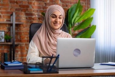Orta Doğulu bej tesettürlü Müslüman kadın çatı katında masa başında çalışıyor. Kadın laptopuyla çalışıyor ve bir şeyler düşünüyor..