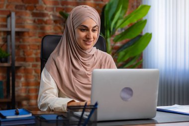 Orta Doğulu bej tesettürlü Müslüman kadın çatı katında masa başında çalışıyor. Kadın laptopuyla çalışıyor ve bir şeyler düşünüyor..