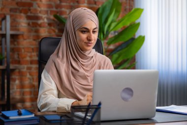 Orta Doğulu bej tesettürlü Müslüman kadın çatı katında masa başında çalışıyor. Kadın laptopuyla çalışıyor ve bir şeyler düşünüyor..