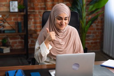 Orta Doğulu bej tesettürlü Müslüman kadın çatı katında masa başında çalışıyor. Kadın dizüstü bilgisayarına umutla bakar ve iyi haberlere ya da galibiyete sevinir..