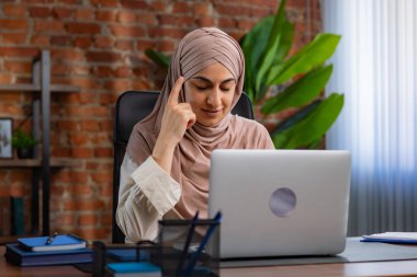 Orta Doğulu bej tesettürlü Müslüman kadın çatı katında masa başında çalışıyor. Kadın laptopuyla çalışıyor ve bir şeyler düşünüyor..