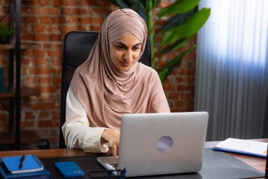 Orta Doğulu bej tesettürlü Müslüman kadın çatı katında masa başında çalışıyor. Kadın dizüstü bilgisayarda klavye kullanıyor..