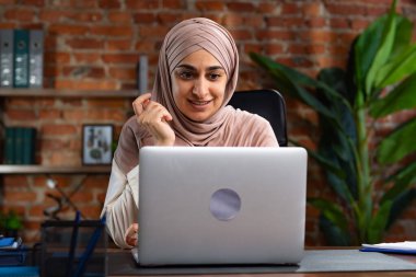Orta Doğulu bej tesettürlü Müslüman kadın çatı katında masa başında çalışıyor. Kadın laptopuyla çalışıyor ve bir şeyler düşünüyor..