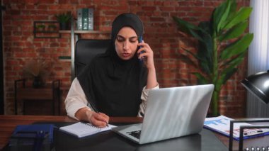Orta Doğulu siyah tesettürlü Müslüman kadın çatı katında masa başında çalışıyor. Kadın telefonda konuşuyor ve bir deftere bilgi yazıyor..