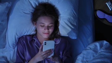 Genç bir kadın gece pijamalarıyla yatakta uzanıyor, akıllı telefon ekranının mavi ışığıyla aydınlatılıyor. Karanlık yatak odası ve parlayan ekran uykusuzluk ve ekran bağımlılığını vurguluyor.