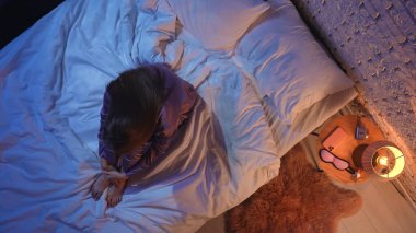 Yatakta kıvrılmış, uykusuzlukla boğuşan saten pijamalı genç bir kadının en iyi görüntüsü. Loş bir komodin lambası ve uyku maskesi gece ayarlarını vurgular.