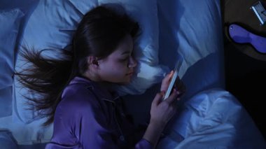 Genç bir kadın gece pijamalarıyla yatakta uzanıyor, akıllı telefon ekranının mavi ışığıyla aydınlatılıyor. Karanlık yatak odası ve parlayan ekran uykusuzluk ve ekran bağımlılığını vurguluyor.