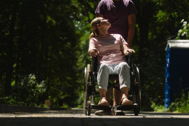 Tekerlekli sandalyede oturan birine, yemyeşil bir parkta güneşli bir yürüyüş yapan başka biri eşlik eder..