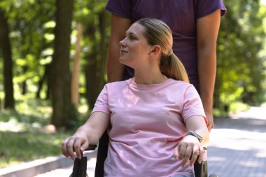 Pembe tişörtlü bir kadın, tekerlekli sandalyede oturuyor. Parkta güneşli bir günün tadını çıkarıyor. Yanında onu iten biri var..