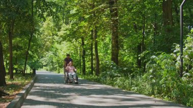 Bir adam tekerlekli sandalyedeki bir kadını bereketli, ağaç çizgili bir patikaya itiyor, doğadaki ulaşılabilirliği vurguluyor..
