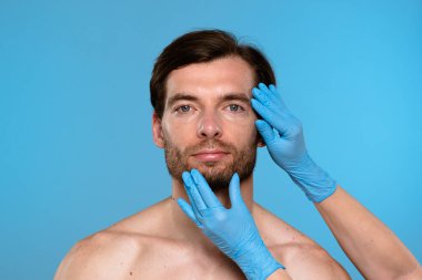 Mavi eldiven giyen bir kozmetolog estetik geliştirme prosedürleri için erkek hastaları muayene ediyor. Klinik ortamdaki erkekler için cilt bakımı ve güzellik danışmanlığı.