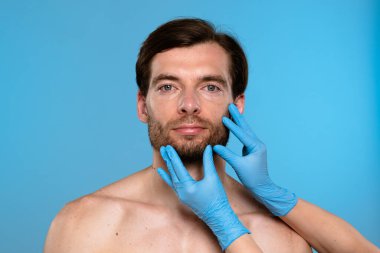Mavi eldiven giyen bir kozmetolog estetik geliştirme prosedürleri için erkek hastaları muayene ediyor. Klinik ortamdaki erkekler için cilt bakımı ve güzellik danışmanlığı.