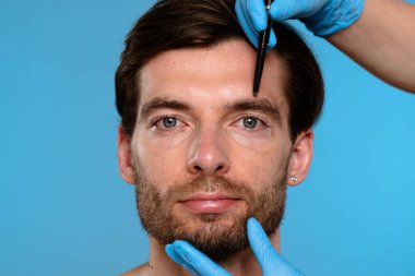 Kozmetik ameliyattan önce yüzü bir tıp uzmanı tarafından işaretlenmiş mavi eldiven takan bir adama yakın çekim. Estetik cerrahi, dermatoloji ya da erkek derisi ile ilgili kampanyalar için uygundur..