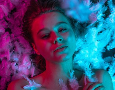 Beyaz tüylerle kaplı, neon pembe ve mavi ışıkla aydınlatılmış bir yüzeyde yatan genç bir güzellik modelinin üst görüntüsü. Güzellik reklamları, cilt tanıtımları, sağlık kampanyaları ya da editör kullanımı için mükemmel..