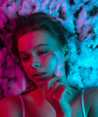 Beyaz tüylerle kaplı, neon pembe ve mavi ışıkla aydınlatılmış bir yüzeyde yatan genç bir güzellik modelinin üst görüntüsü. Güzellik reklamları, cilt tanıtımları, sağlık kampanyaları ya da editör kullanımı için mükemmel..