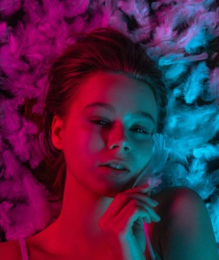 Beyaz tüylerle kaplı, neon pembe ve mavi ışıkla aydınlatılmış bir yüzeyde yatan genç bir güzellik modelinin üst görüntüsü. Güzellik reklamları, cilt tanıtımları, sağlık kampanyaları ya da editör kullanımı için mükemmel..