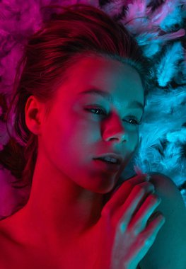 Beyaz tüylerle kaplı, neon pembe ve mavi ışıkla aydınlatılmış bir yüzeyde yatan genç bir güzellik modelinin üst görüntüsü. Güzellik reklamları, cilt tanıtımları, sağlık kampanyaları ya da editör kullanımı için mükemmel..