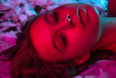 Neon pembe ve mavi ışıkta beyaz tüylerin üzerinde uzanan ışıl ışıl genç bir kadının üst görüntüsü. Güzellik içeriği, güzellik endüstrisinde reklam kampanyaları ve sağlık sektöründe markalaşma için mükemmel..