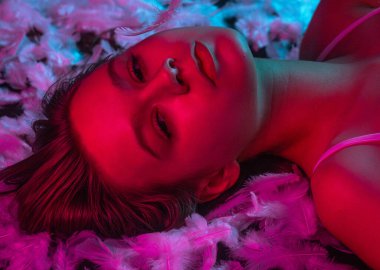 Neon pembe ve mavi ışıkta beyaz tüylerin üzerinde uzanan ışıl ışıl genç bir kadının üst görüntüsü. Güzellik içeriği, güzellik endüstrisinde reklam kampanyaları ve sağlık sektöründe markalaşma için mükemmel..