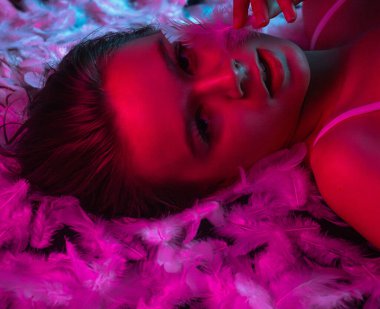Neon pembe ve mavi ışıkta beyaz tüylerin üzerinde uzanan ışıl ışıl genç bir kadının üst görüntüsü. Güzellik içeriği, güzellik endüstrisinde reklam kampanyaları ve sağlık sektöründe markalaşma için mükemmel..