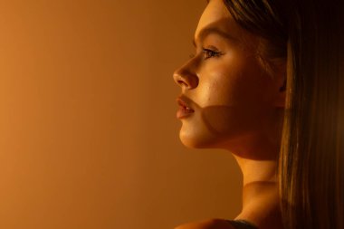 Yüzünde geometrik gölgeler olan genç bir kadının yumuşak turuncu arka planına yakın çekim. Güzellik, cilt bakımı ve duygusal fotoğrafçılık konuları için mükemmel..
