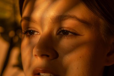 Yüzünde geometrik gölgeler olan genç bir kadının yumuşak turuncu arka planına yakın çekim. Güzellik, cilt bakımı ve duygusal fotoğrafçılık konuları için mükemmel..