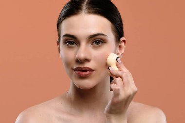 Bir kadın doğal görünmek için sünger kullanarak makyaj yapıyor. Süngeri yanağında tutuyor..