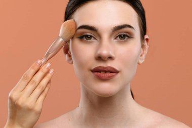 Genç bir kadın yüzüne fırça sürerek doğal ve kusursuz bir görünüm elde ederek makyaj yapar. Arka plan şeftali renginde, sahnenin zarafetini arttırıyor..