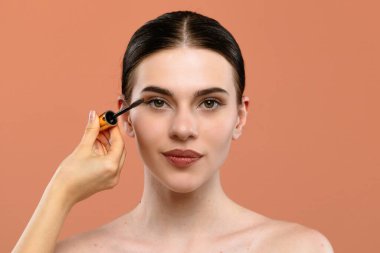 Bir kadın göz makyajını geliştirmek için rimel sürüyor. Şeftali arka planına güzellik ve tarz vurguluyor..