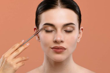 Gözleri kapalı bir kadın şeftali arkaplanda fırça kullanarak göz makyajı yaptırıyor..