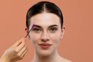 Kaşları bakımlı bir kadın şeftali arka planında mor renkli bir fırça ile kaşlarını fırçalatıyor..