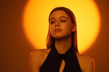 Bir kadın sıcak, dramatik turuncu ışıkta yakalanıyor, özelliklerini vurguluyor ve dengesiz bir atmosfer yaratıyor..