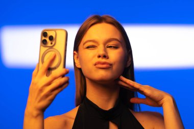 Bir kadın akıllı telefonuyla canlı mavi bir arkaplan kullanarak çağdaş teknoloji ve kişisel ifade sergileyen bir selfie çekiyor..