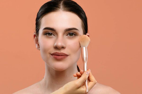 A woman applies makeup with a brush, highlighting clear skin and natural beauty against a peach background.