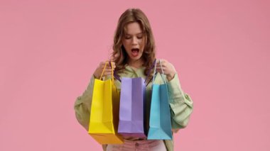 Genç bir kadın pembe bir arka plana renkli alışveriş torbaları tutarken heyecanını ifade ediyor..