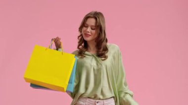 Genç bir kadın alışverişi seviyor, pembe arka planda parlak renkli çantalar tutuyor, açık yeşil bir bluz ve beyaz pantolon giyiyor..