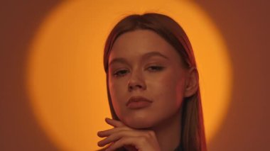 Kadın kameraya bakar ve yanağını çarpıcı bir ışıkla parlak turuncu bir arka plana vurur.