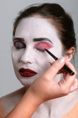 Sahne makyajı yaptıran bir kişi beyaz tabanlı, kırmızı gözlü ve koyu siyah kaşlı..